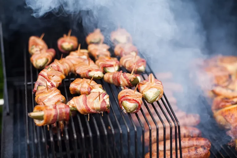 Hasty Bake jalapeño poppers wrapped in bacon grilling on the grates with smoke, on a Hasty Bake Charcoal Grill