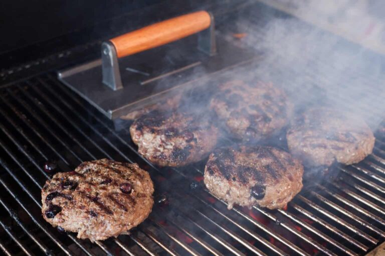 Freshly ground venison burger patties grilling on the grates of a Hasty Bake Charcoal Grill with smoke rising