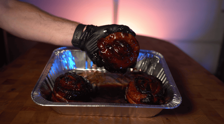 Gloved hand picking up a glossy smoked pork belly cinnamon roll from a foil tray on a Hasty Bake Charcoal Grill